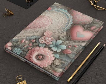 May include: A pink and gray floral patterned journal with a lace design and hearts. The journal is closed and lying on a gray surface.