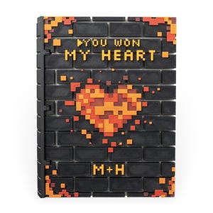 Pode incluir: Uma parede de tijolo preto com um coração pixelado em laranja e vermelho. O texto "YOU WON MY HEART" está em letras amarelas na parede. O texto "M+H" está em letras amarelas na parte inferior da parede.