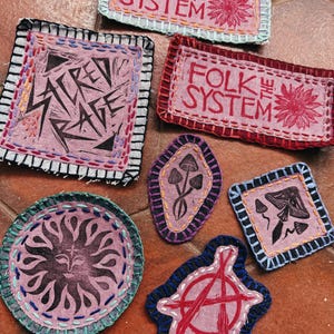 May include: A collection of handmade fabric patches with various designs and text. The patches feature embroidered edges and include phrases like "FOLK THE SYSTEM" and "STREET RAGE." Other patches display a sun, mushrooms, and an anarchy symbol.