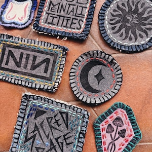 May include: A collection of handmade fabric patches with embroidered designs and text. The patches feature various shapes, including squares, circles, and a diamond. Designs include a sun, a crescent moon, mushrooms, and the text "MOB MENTALITIES" and "NIN".