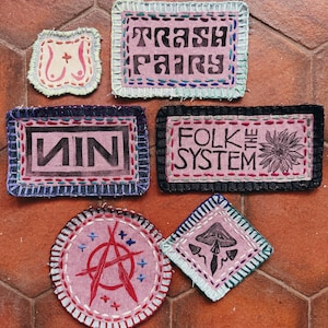 May include: A collection of handmade fabric patches with embroidered edges. The patches feature various designs, including text that reads "TRASH FAIRY", "MIN", and "FOLK THE SYSTEM". Other patches display a mushroom, a flower, and an anarchy symbol.