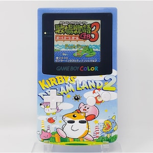 Może przedstawiać: Niebieska przenośna konsola do gier Game Boy Color z włożoną grą Kirby's Dream Land 2. Konsola ma kolorowy design z postaciami Kirby'ego i tekstem "Kirby's Dream Land 2". Ekran wyświetla grę z serii "牧場物語", z widocznym tekstem "Push Start".
