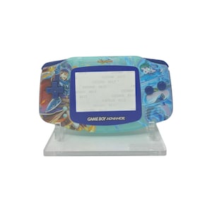 Puede incluir: Una consola portátil Game Boy Advance con un diseño azul y turquesa. La consola presenta un borde de pantalla azul y un soporte de plástico transparente. El texto "GAME BOY ADVANCE" es visible en la parte inferior.