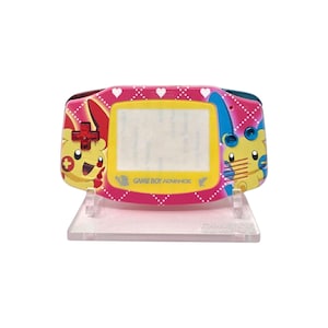 Könnte beinhalten: Ein pinkfarbener Game Boy Advance Handheld-Spielkonsole mit Pikachu-Design. Die Konsole verfügt über einen Bildschirm, Tasten und ein Steuerkreuz. Das Design umfasst Herzen und die Worte "Game Boy Advance". Die Konsole steht auf einem transparenten Ständer.