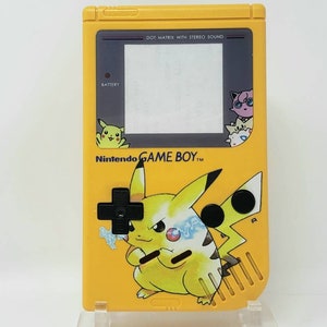 Peut inclure: Console de jeu vidéo portable Game Boy Color jaune avec un design Pikachu. La console a un écran noir et blanc, un pavé directionnel noir et des boutons noirs. Le texte "Nintendo GAME BOY" est imprimé sur la face avant de la console.