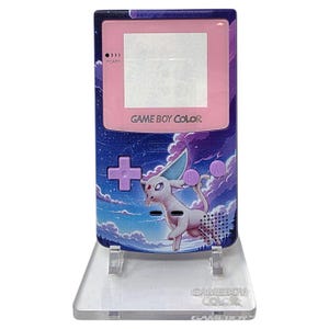 Op de afbeelding: Een Game Boy Color handheld console met een aangepast ontwerp met een paarse en blauwe sterrenhemel en een wit en roze cartoonkarakter. De console wordt gepresenteerd op een transparante acryl standaard.