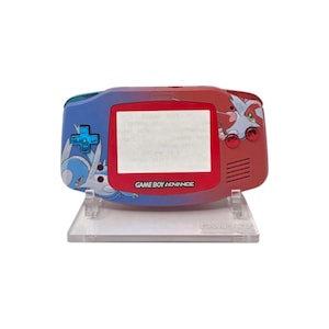 Puede incluir: Una consola portátil Game Boy Advance personalizada. El dispositivo presenta un diseño degradado azul y rojo con ilustraciones de personajes. La pantalla está enmarcada en rojo y el texto "GAME BOY ADVANCE" es visible debajo.