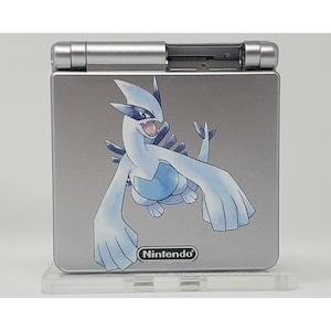 Puede incluir: Consola de juegos portátil Nintendo Game Boy Advance SP plateada con un gráfico de Lugia. La consola tiene una bisagra plateada y un logotipo de Nintendo. El gráfico de Lugia es azul y blanco.