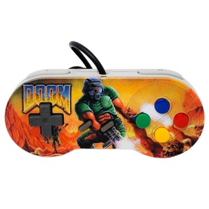 May include: A white Super Nintendo controller with a custom design featuring the Doom video game logo and a character from the game. The controller has a black cord and four colored buttons.