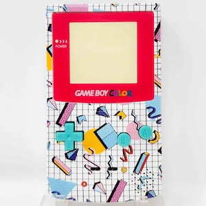 Könnte beinhalten: Eine weiße Game Boy Color Handheld-Videospielkonsole mit einem rosa Bildschirm und einem bunten geometrischen Muster aus den 90er Jahren auf der Vorderseite. Die Knöpfe sind türkis, gelb und rosa.