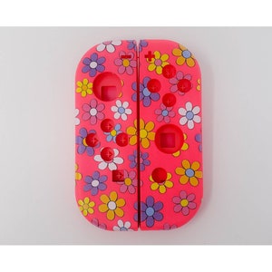 Peut inclure: Paire de manettes Joy-Con roses pour Switch avec un motif floral. Les manettes présentent un motif répété de fleurs blanches, jaunes, bleues et violettes. Les boutons et les joysticks sont visibles.