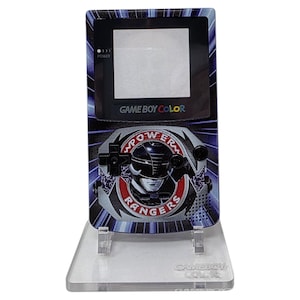 Op de afbeelding: Een Game Boy Color handheld console met een Power Rangers ontwerp. De console heeft een zwart scherm en knoppen, met een afbeelding van een Power Ranger helm en de woorden "Power Rangers" in een rood en wit embleem. De achtergrond is blauw en zwart.