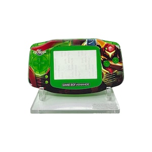 May include: A custom Game Boy Advance handheld console featuring a Metroid theme. The device has a green and red color scheme with artwork of the character Samus Aran. The screen is surrounded by a green bezel, and the text "GAME BOY ADVANCE" is visible.