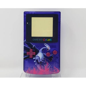 May include: A purple Game Boy Color handheld video game console with a translucent purple shell. The console features a  "GAME BOY COLOR" logo and a graphic of a white and blue Pokemon character on a pink and purple background.