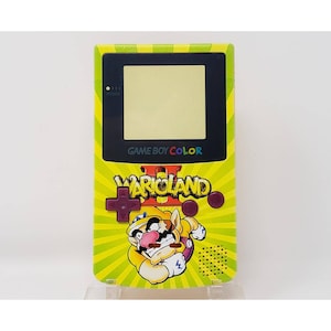 Op de afbeelding: Een gele Game Boy Color draagbare videogameconsole met een Wario Land II gamecartridge erin. De console heeft een geel en groen ontwerp met een cartoonafbeelding van Wario aan de voorkant.
