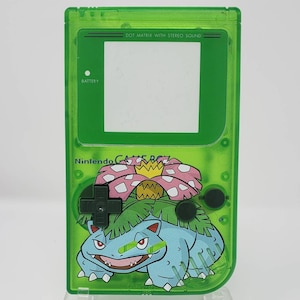 May include: A green Game Boy Color with a clear plastic shell. The front of the Game Boy Color features a  'DOT MATRIX WITH STEREO SOUND' screen and a  'BATTERY' label. The Game Boy Color has a  'Nintendo GAME BOY' logo and a graphic of a green, blue, and pink Pokemon character.