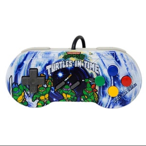 May include: A white Teenage Mutant Ninja Turtles video game controller with a blue and black background featuring the Teenage Mutant Ninja Turtles characters. The controller has a black directional pad, four gray buttons, and four colored buttons. The text "Teenage Mutant Ninja Turtles" and "Turtles in Time" are printed on the controller.