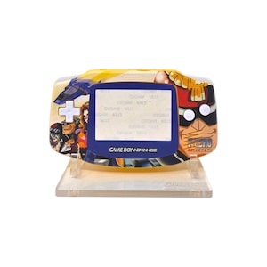 May include: A custom-painted Game Boy Advance handheld console. The device features a colorful design with anime-style characters and the text "ZERO LEGEND". The screen is blue, and the buttons are white. It is displayed on a clear stand.
