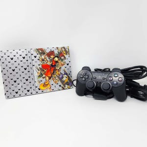 May include: A black Sony PlayStation 2 console with a Kingdom Hearts game case. The case has a silver background with a pattern of crowns and Mickey Mouse heads. The front of the case features a colorful illustration of Sora, the main character from the game, along with other characters. The console is also accompanied by a black PlayStation 2 controller.