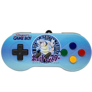 May include: A blue Super Game Boy controller with a Pokemon design. The controller features a gray plus button, four colored buttons, and a circular image of a Pokemon character with the text "Pocket Monsters" in Japanese.