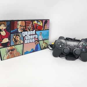 May include: A black PlayStation 2 console with a custom Grand Theft Auto design. The console features a collage of characters and scenes from the game. A black PlayStation 2 controller is also included.