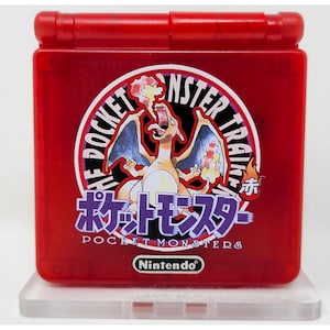 Op de afbeelding: Rode Game Boy Advance SP-console met een ronde sticker. De sticker toont een Charizard-illustratie en Japanse tekst. De tekst luidt "POCKET MONSTER TRAINER" en "Nintendo" staat onderaan afgedrukt.