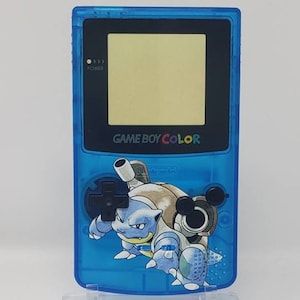 Puede incluir: Una consola de videojuegos portátil Game Boy Color azul con una carcasa azul transparente y una pantalla negra. La consola presenta un gráfico del Pokémon Squirtle en la parte delantera.
