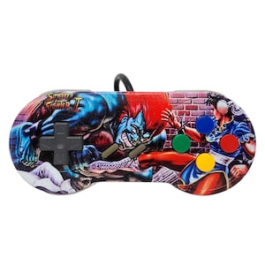 May include: A white Super Nintendo controller with a custom design featuring a red-haired, blue-skinned character fighting a female character in a blue and red outfit. The design includes the words "Street Fighter II" on the controller.