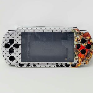 May include: A silver and black PlayStation Portable (PSP) with a Kingdom Hearts design. The design features characters from the game, including Sora, Riku, and Kairi. The PSP has a black and white pattern on the front and back.