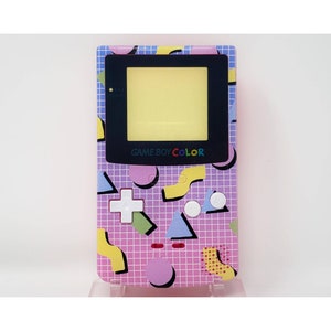 Puede incluir: Una consola de videojuegos portátil Game Boy Color rosa con un diseño de patrón geométrico. La consola tiene una pantalla negra y botones blancos.