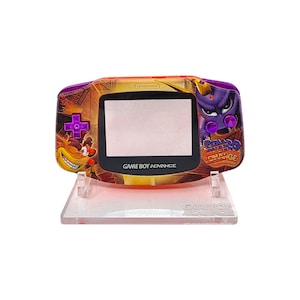 May include: A custom-designed Game Boy Advance handheld console. The device features a purple and orange shell with a Crash Bandicoot and Spyro the Dragon theme. The screen is black, and the buttons are purple. The console is displayed on a clear stand.