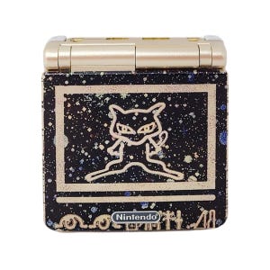 May include: A gold Game Boy Advance SP with a black and silver design featuring a stylised image of a black cat with white eyes. The text "Nintendo" is visible on the bottom of the device.