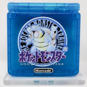 May include: A blue Game Boy Color handheld video game console with a clear plastic shell. The console features a circular sticker with a cartoon image of a blue and yellow turtle-like creature. The sticker also includes the text "The Pokemon Trainer" and "Pocket Monsters" in Japanese and English. The Nintendo logo is visible on the bottom of the console.