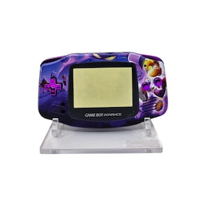 May include: A purple Game Boy Advance handheld console with a Rayman-themed design. The console features a black screen and purple buttons. The text "GAME BOY ADVANCE" is printed below the screen. It is displayed on a clear plastic stand.