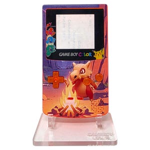 Op de afbeelding: Aangepaste Game Boy Color-console met een Cubone-personageontwerp en een kampvuurscène. De console staat op een transparante standaard. De woorden "GAME BOY COLOR" zijn zichtbaar.