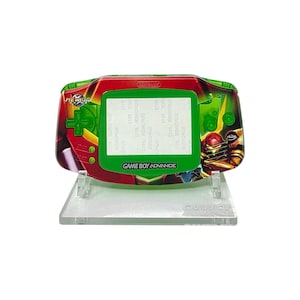 May include: A custom Game Boy Advance console featuring a Metroid design. The handheld console has a green screen and a red, green, and black exterior with the words "Metroid" and "Game Boy Advance" visible. It is displayed on a clear stand.