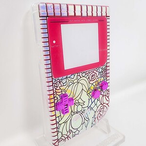 Puede incluir: Una Game Boy Color rosa y blanca con un diseño floral y botones morados. La pantalla dice "DOT MATRIX WITH STEREO SOUND" y "BATTERY".