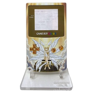 Puede incluir: Consola de juegos portátil Game Boy Color dorada con un diseño personalizado de ángel. Se muestra en un soporte de plástico transparente. La pantalla muestra texto. Los botones son dorados.