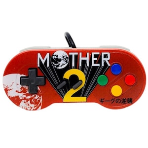 May include: A red and white Super Nintendo controller with a black and yellow "MOTHER 2" logo and a black and white image of a planet. The controller has four colored buttons and the text "キーダの逆襲" in Japanese.