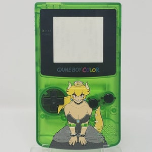 Könnte beinhalten: Ein transparentes grünes Game Boy Color Handheld-Konsolen-Gerät mit einem Bowser-Themen-Grafik. Die Konsole verfügt über einen schwarzen Bildschirm, Tasten und den Text "GAME BOY COLOR". Die Grafik zeigt eine Zeichentrickfigur mit Krone und schwarzem Kleid.