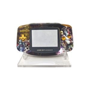 Könnte beinhalten: Ein schwarzer Game Boy Advance Handheld-Konsolen mit einem individuellen Golden Sun-Design. Die Konsole verfügt über einen Bildschirm, Steuertasten und den Text "GAME BOY ADVANCE". Das Design umfasst Charaktere und das Golden Sun-Logo.