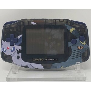 Puede incluir: Una Game Boy Advance transparente azul con un diseño de Pokemon. El diseño presenta un Eevee blanco y azul a la izquierda y un Espeon azul y amarillo a la derecha. La Game Boy Advance está en un soporte transparente.