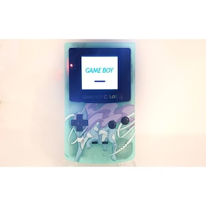 Op de afbeelding: Een turquoise Game Boy Color handheld-spelconsole met een aangepast ontwerp met een blauw en paars Pokémon-personage. Het scherm toont de tekst "GAME BOY" in een blauw lettertype.