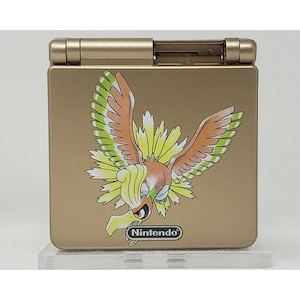 Op de afbeelding: Goudkleurige Nintendo Game Boy Advance SP handheld gameconsole. De console heeft een kleurrijke afbeelding van een vogelachtig wezen met uitgespreide vleugels. Het Nintendo-logo is onderaan zichtbaar.
