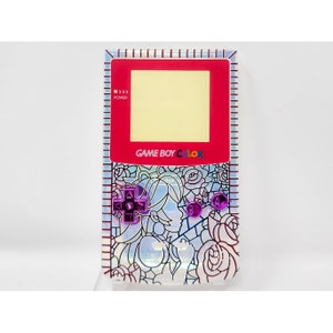 May include: A red Game Boy Color with a clear, iridescent shell. The shell features a floral design with a girl's face in the center. The buttons are purple.