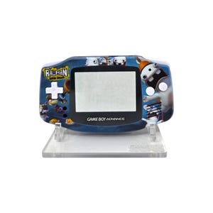 Puede incluir: Consola de juegos portátil Game Boy Advance azul con diseño "Rayman Raving Rabbids". La consola tiene una pantalla, botones y el texto "Game Boy Advance". Se muestra en un soporte de plástico transparente.