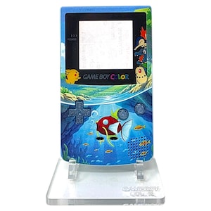May include: A blue Game Boy Color handheld game console with a custom design featuring underwater scenes and Pokémon characters. The console has a clear stand and the text "GAME BOY COLOR" is visible.
