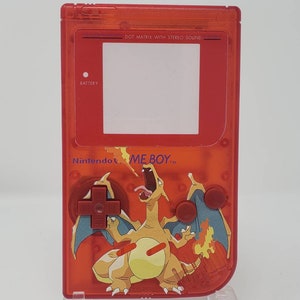 May include: A red Game Boy Color handheld video game console with a clear plastic shell. The console features a Pokemon character, Charizard, on the front. The text "Nintendo" and "GAME BOY" are visible on the console.