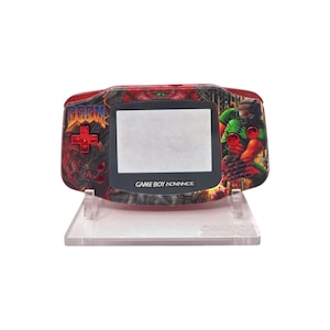 May include: A red and black Game Boy Advance handheld console with a custom DOOM design. The console features a D-pad and the text "GAME BOY ADVANCE". The design includes a green and red character graphic.