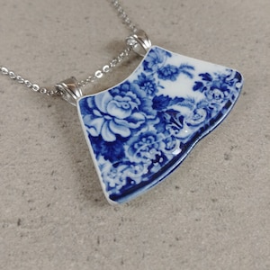 以下が含まれることがあります： 青い花柄の磁器製のペンダントネックレス。ペンダントは扇形をしていて、銀色のチェーンに繋がっています。